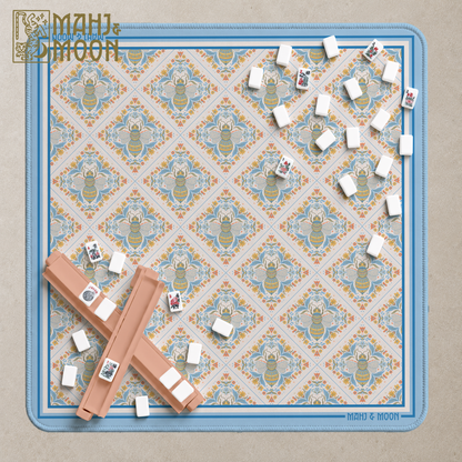 A top-down view of a Mahjong game set by MAHJ & MOON, featuring a square playmat with a blue border and a repeating beige, blue, and green floral bee pattern. The playmat is styled with several tiles arranged on it and two tan tile racks in the lower-left corner, set against a neutral textured beige background.