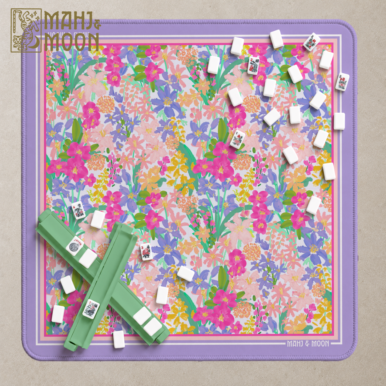 A Mahj & Moon mahjong game set displayed on a square floral mat with a lavender border. The mat features a dense pink, purple, yellow, and green floral pattern, with several green racks holding white-backed tiles and a few loose tiles scattered around.