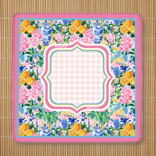 Colorful floral chinoiserie patterned Mahjong mat with bright pink border and light gingham play center shown paced flat on a bamboo background.